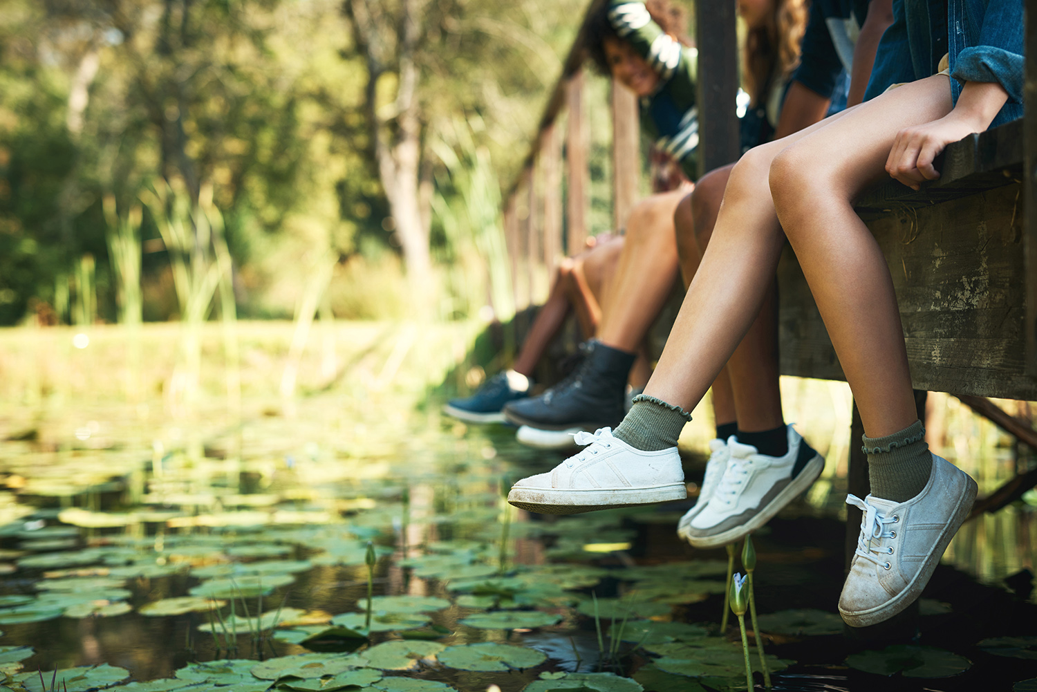 group of diverse smiling teens sitting on a wooden bridge over a pond swinging their legs.